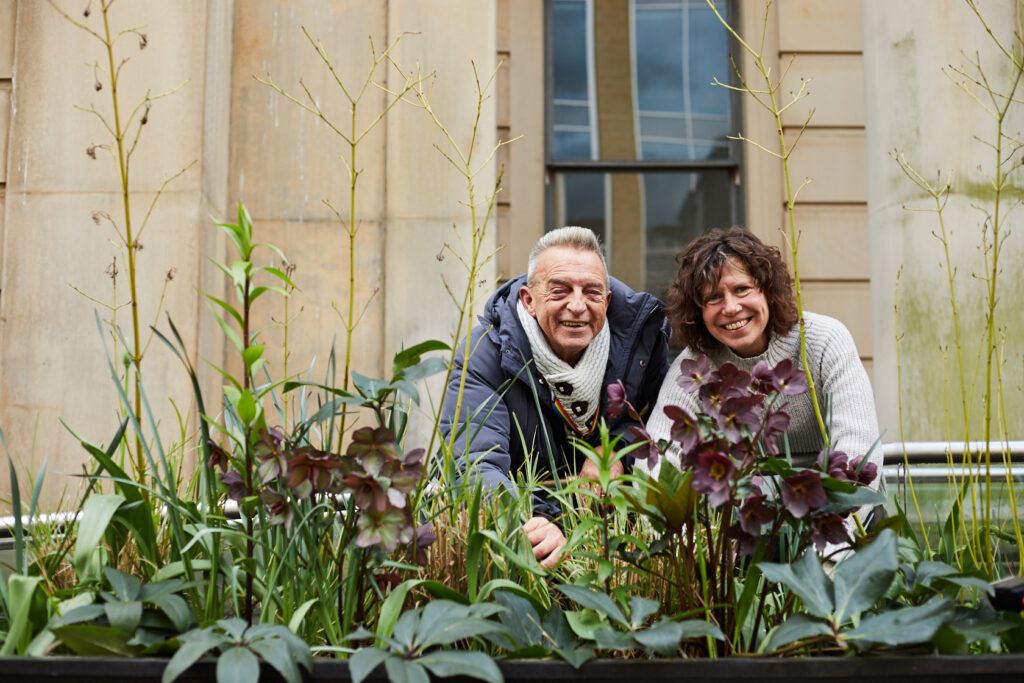 Two older people in a garden, smiling behind plants.