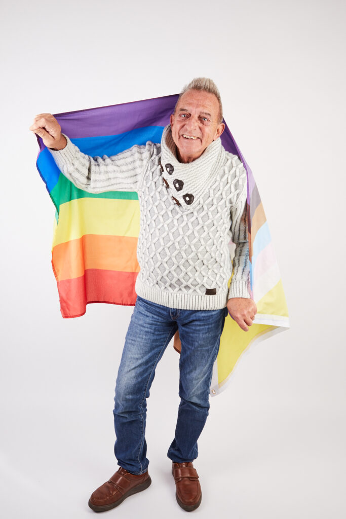 An older man smiling to camera, covered by Pride flag. Portrait.