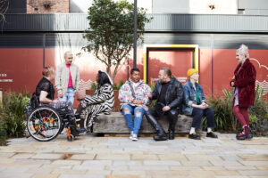 People of various genders, ethnicities and physical states talking to each other in small groups around a wooden bench. Three people on left side , two in the middle, and two on the right.