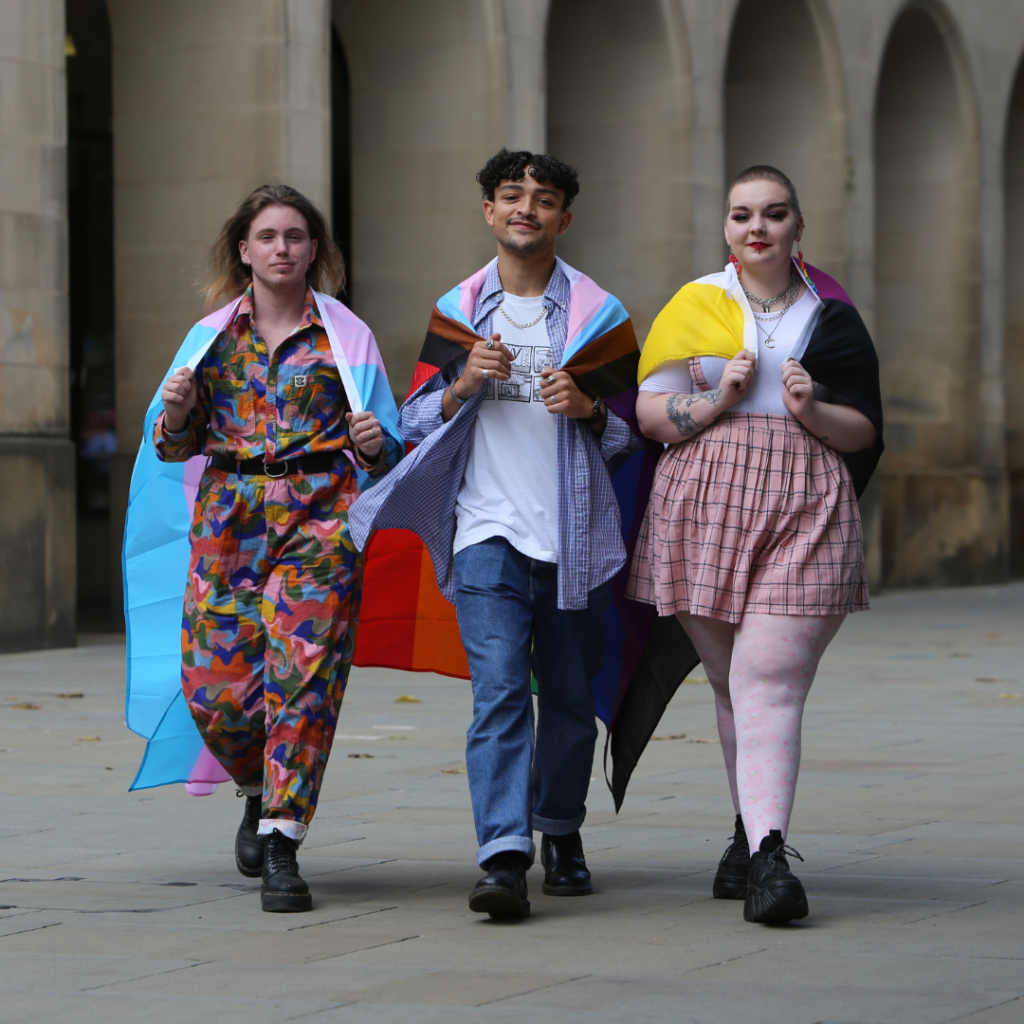 Three people walking confidently towards camera. Masc-presenting person with long hair on the left covered with huge trans flag, brown man with short curly hair in the middle covered with huge rainbow flag, and fem-presenting person with short hair on the right covered with huge non-binary flag.
