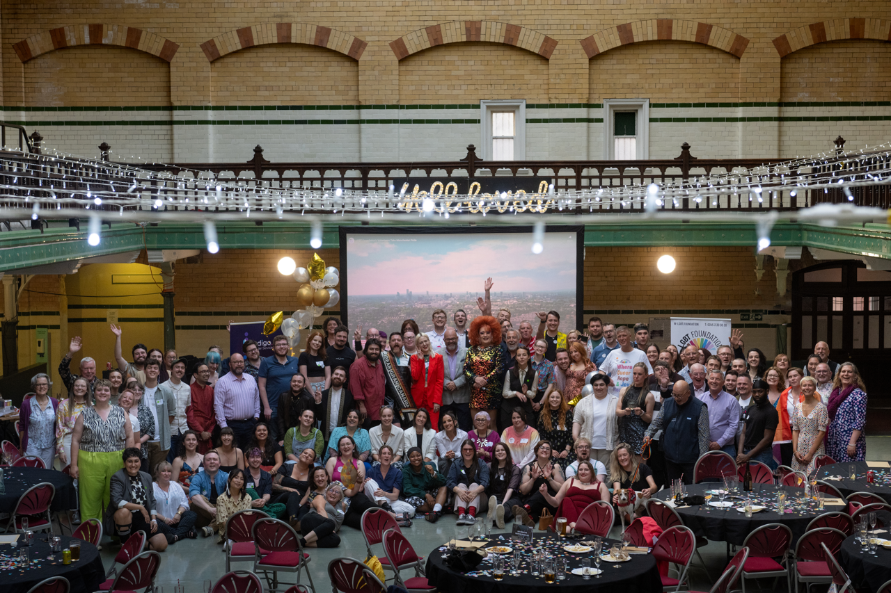 A large group of over a hundred people cheering and smiling at camera. Drag queen with red hair in middle. Background of the venue of Victoria Bath.