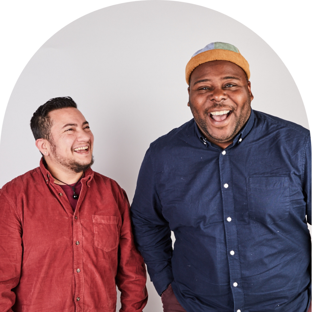 Two masc-presenting people laughing. One looking at the other. Portrait.