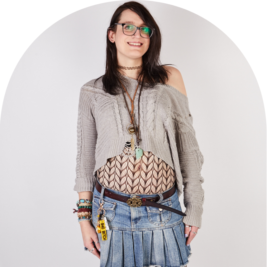 White trans woman in grey jumper and short denim skirt smiling at camera. Hands on thighs. Portrait.