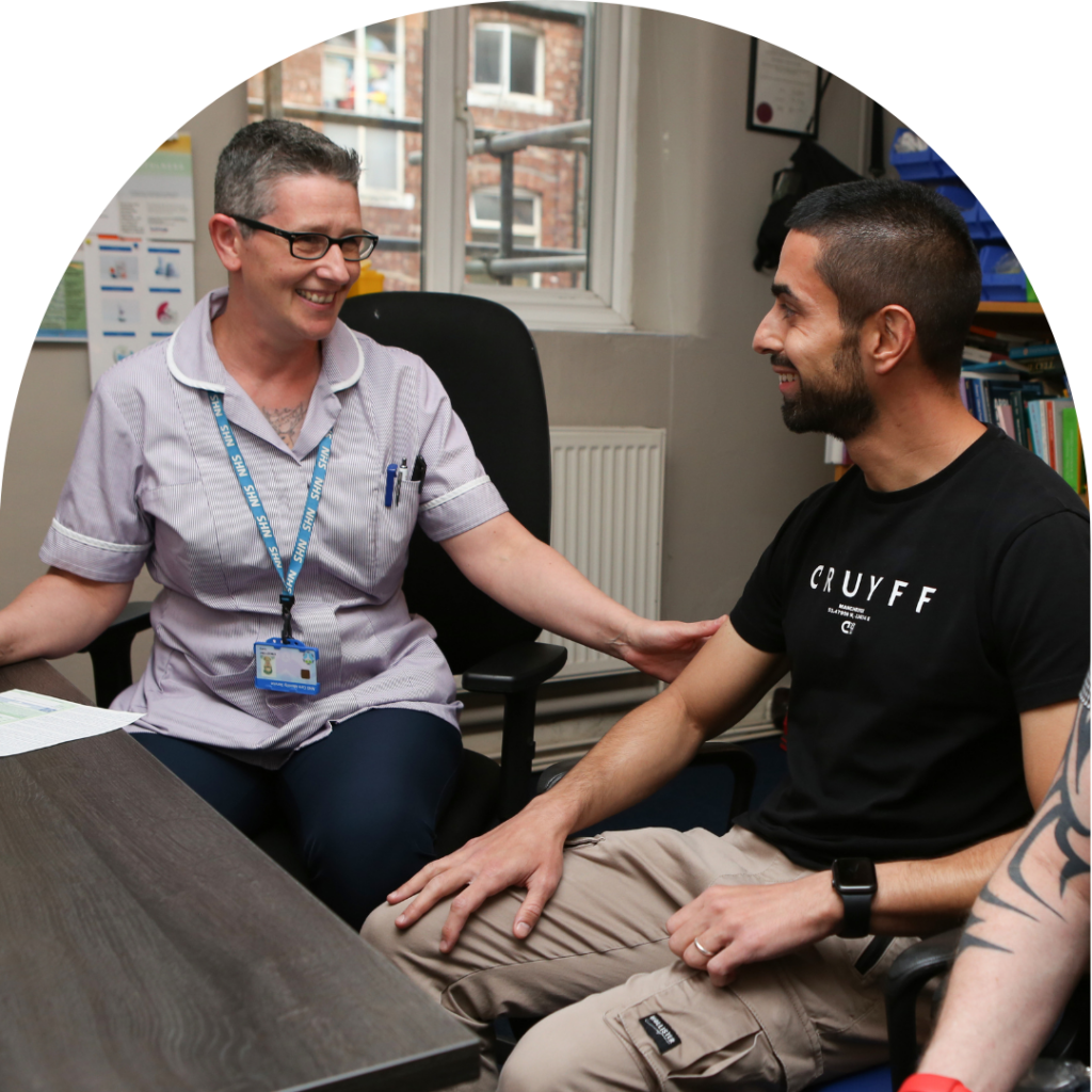 Older female NHS doctor talking to younger brown man in clinic room, giving advice. Both smiling. Focus on doctor.