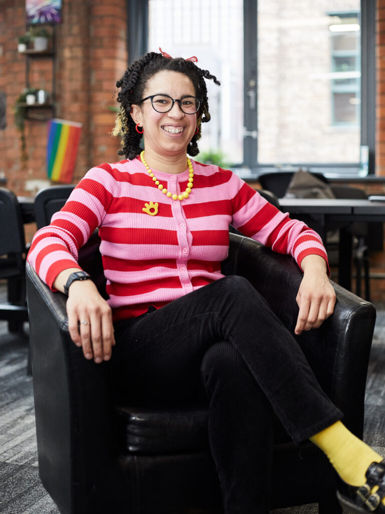 Woman in striped red and pink shirt sitting in sofa chair, legs crossed, smiling at camera.
