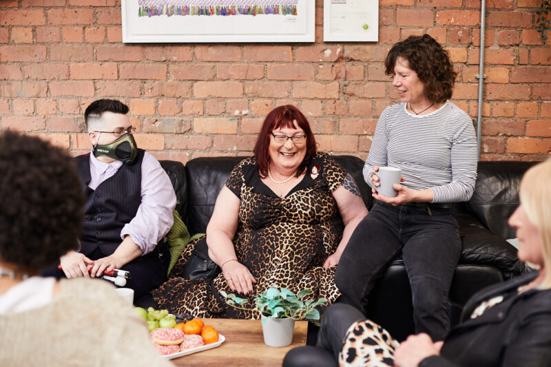 Two fem-presenting and one masc-presenting people sitting on sofa with a table in the front. All smiling and listening to a person outside the frame talking. Table full of fruit, snacks and coffee cups.