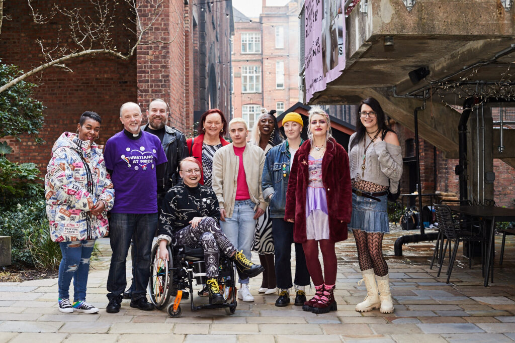 A group of diverse people standing in an alleyway, smiling at camera. In the middle, a fem-presenting person sitting on wheelchair. On the side, people of different gender identities standing in a line. Group portrait.