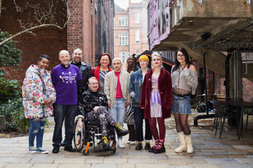 A group of diverse people standing in an alleyway, smiling at camera. In the middle, a fem-presenting person sitting on wheelchair. On the side, people of different gender identities standing in a line. Group portrait.