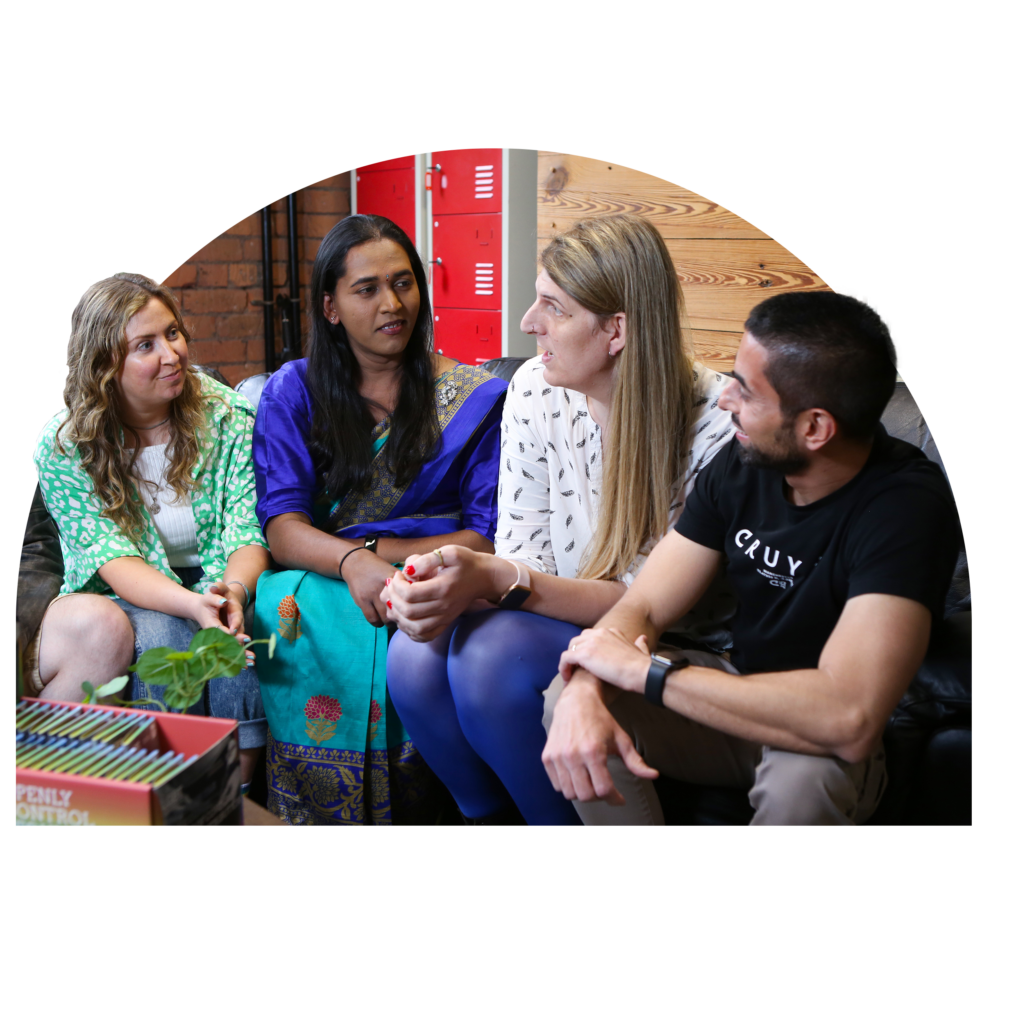 Four people chatting on a sofa. From far left side, white woman in floral top, Asian trans woman in traditional outfit, white trans woman in white blouse, and Asian man in black t-shirt listening carefully, interested.
