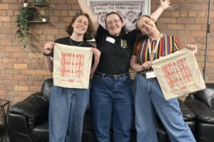 Three woman standing in front of brick wall at LGBT Foundation hub, holding cloths with 'Sugar and Spice' and 'LGBT foundation' on it. All laughing and cheering at camera.