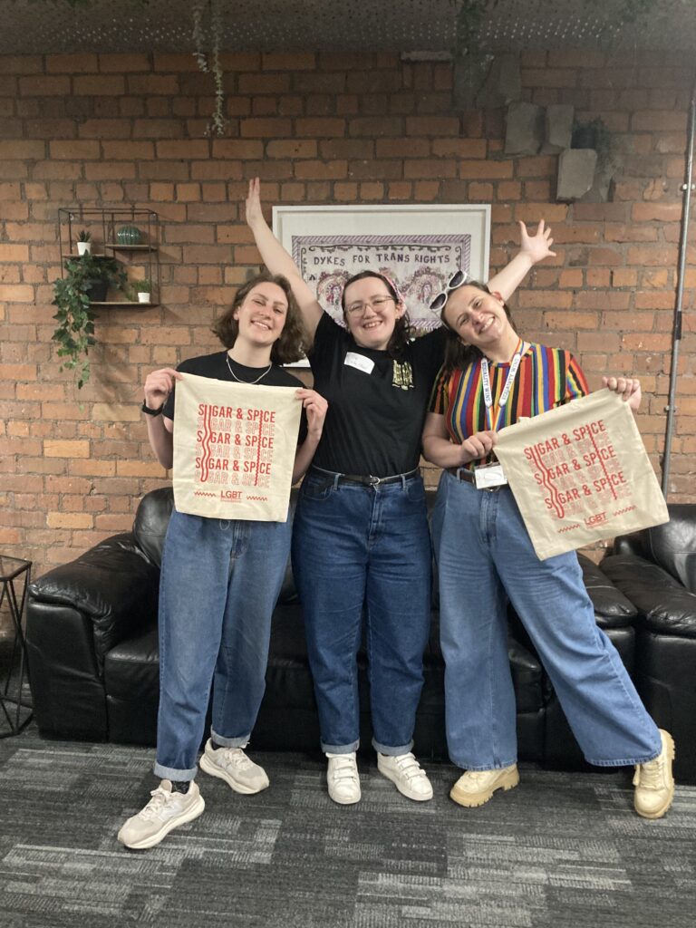 Three woman standing in front of brick wall at LGBT Foundation hub, holding cloths with 'Sugar and Spice' and 'LGBT foundation' on it. All laughing and cheering at camera.