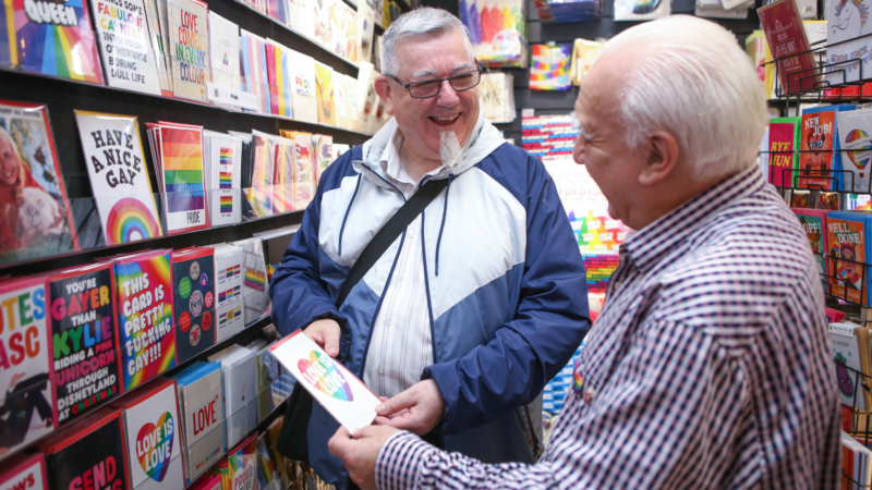 Two older gay men with white hair selecting cards in card shop for Pride. Both looking at each other and smiling.