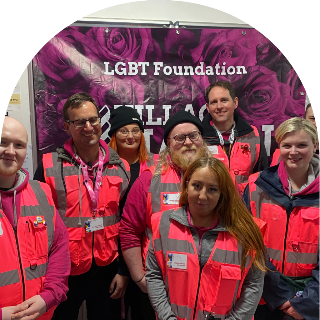 A large group of village angels in pink hi vis vests standing in front of a purple banner reading 'LGBT Foundation Village Haven', smiling at camera. Group portrait.
