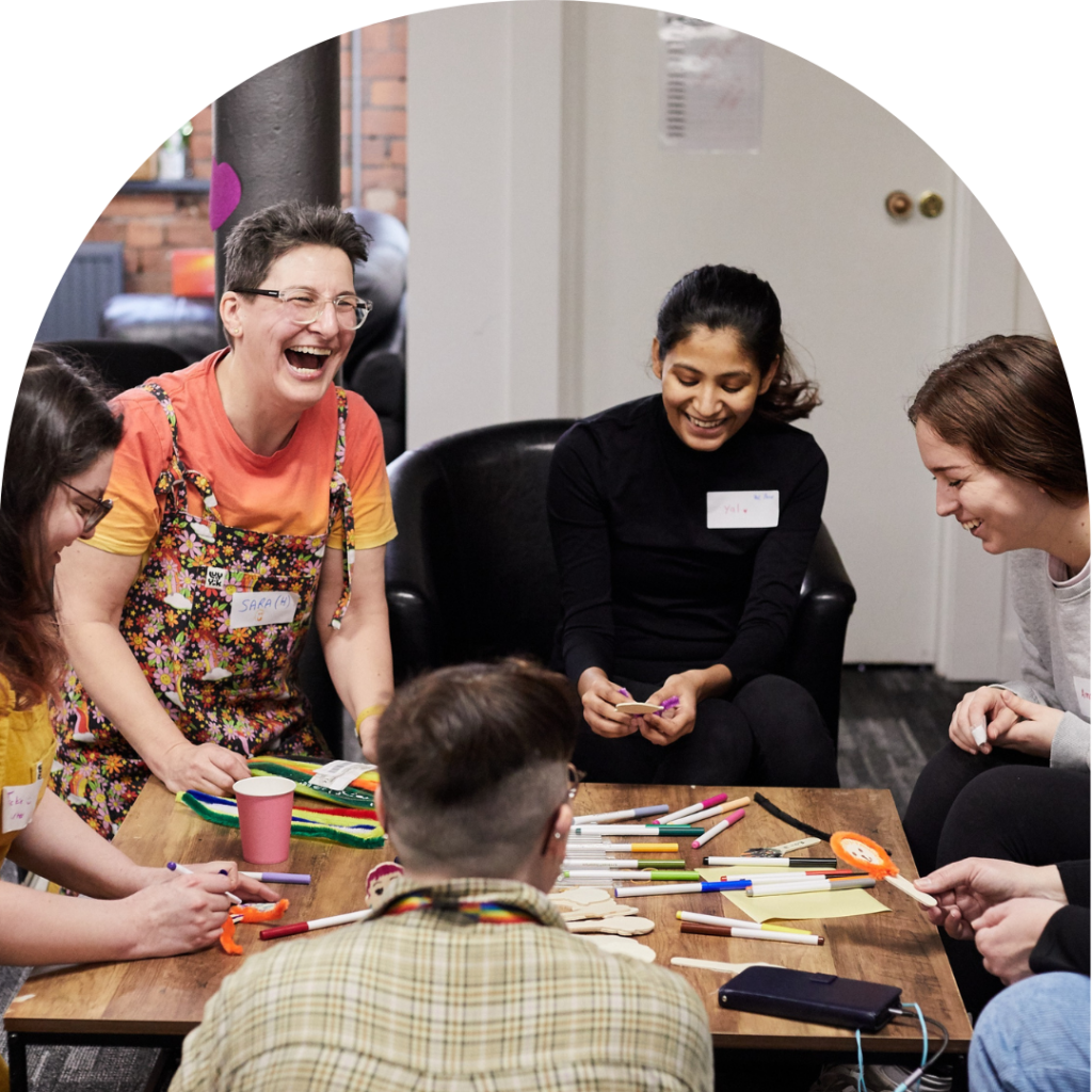 A group of women and non-binary people sitting around a table, doing a workshop. Colouring markers and paper scattered all over the table. All laughing.
