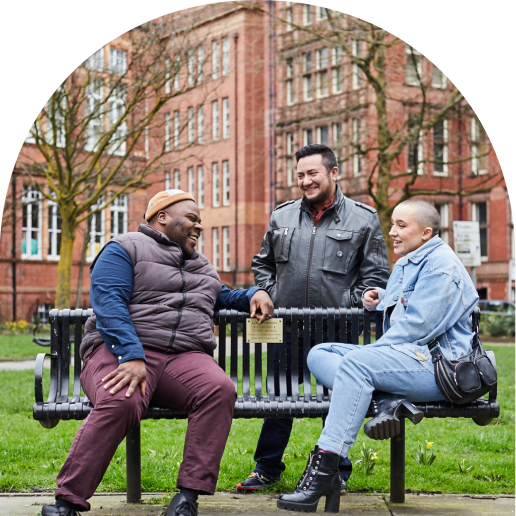 Three friends chatting and smiling around a bench in Sackville gardens.