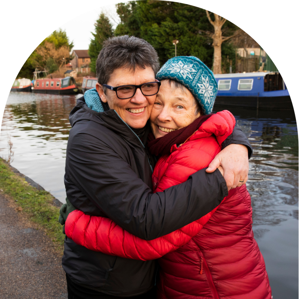 Two older women in winter coats hugging each other tightly next to the canal, smiling at camera. Portrait.