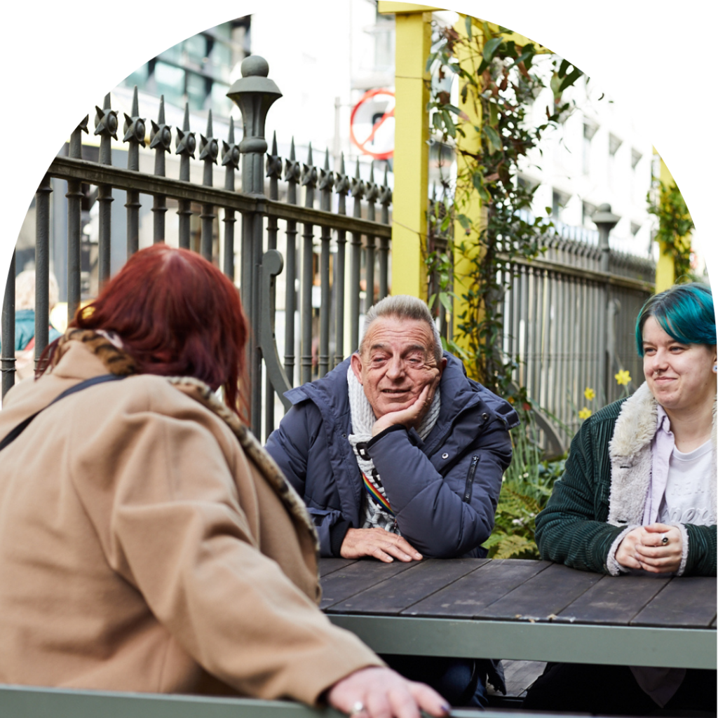 Three people chatting around a table in a garden. An older man and a non-binary person with blue hair facing the camera, smiling. On the opposite sits a trans woman with red hair.