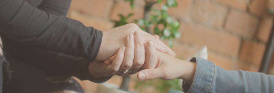 Person on left using both hands to hold hand of the person on right. Person on left wearing a diamond ring on ring finger. Close-up to hands.
