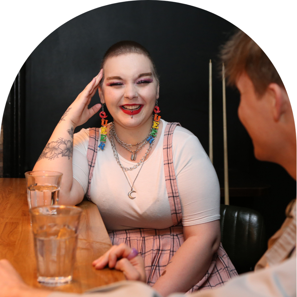 A white person sits laughing, they have a glass of water on a bar in front of them and they are wearing a white t-shirt and pink skirt with matching makeup, their earings spell the word queer. They are with a friend who is masc presenting and wearing a grey jacket