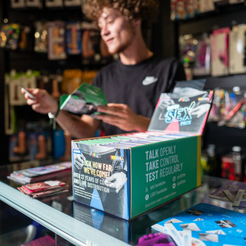 our safer sex packs can be seen on the counter in a shop and the resources are being reviewed by a young trans masc person who is intently reading the information about the condoms and lube inside the packs