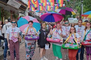 Our sexual health team and their volunteers stand on canal street with their freebies including condoms and lube using their ice cream tray mobile outreach, they're all holding identity based LGBTQ+ flag umbrellas.