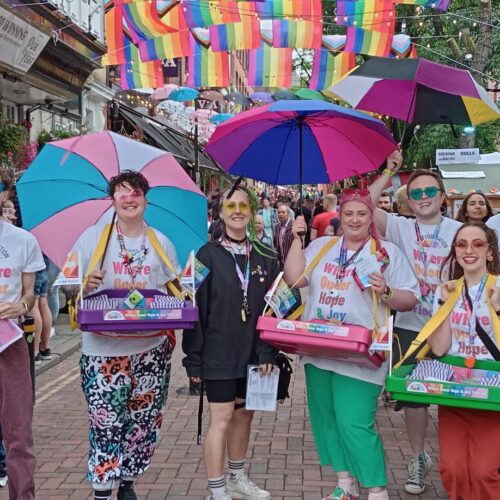 Our sexual health team and their volunteers stand on canal street with their freebies including condoms and lube using their ice cream tray mobile outreach, they're all holding identity based LGBTQ+ flag umbrellas.