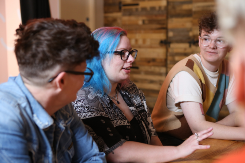 Image shows a group of queer people in discussion around a table